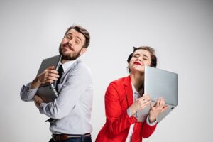 Two people stand with their eyes closed, smiling and hugging their laptops. The person on the left wears a light blue shirt and tie, and the person on the right wears a red blazer, celebrating after upgrading to Office 365.