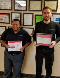 Two technology visionaries wearing black shirts hold certification plaques while standing in front of a wall filled with framed certificates and awards.