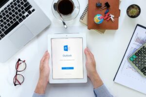 A person holding a tablet displaying the Outlook app login screen, with a laptop, coffee cup, notebooks, small globe, and calculator on a desk.