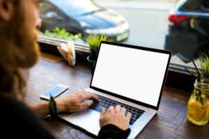 Person using a laptop with a blank screen at a wooden table near a window. A smartphone, plants, napkins, and a jar with a drink are also on the table. Cars can be seen outside the window.