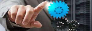 A person in a suit points at a digital, glowing blue gear above several black gears, symbolizing technology and connectivity. The background features blurred machinery or server racks.