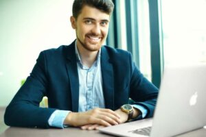 Man in a suit sits at a desk smiling, with his hands resting on the surface next to an open laptop.